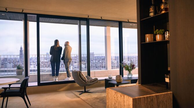 Alt: Vista interna di un appartamento moderno con ampie porte scorrevoli e due persone su un balcone.