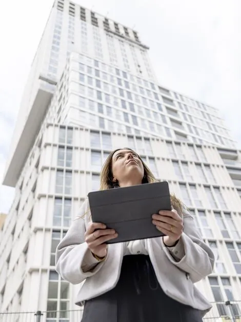 Femme blonde tenant un iPad regardant vers l’immeuble d’appartements blanc.
